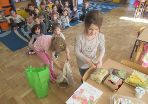 Dziewczynki wyjmują produkty z toreb, pozostałe dzieci siedzą na dywanie.