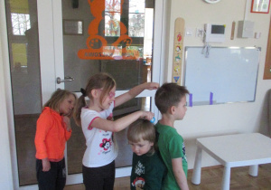 Dwóch chłopców stoi plecami do siebie, dziewczynka sprawdza o ile jeden chłopiec jest wyższy od drugiego.