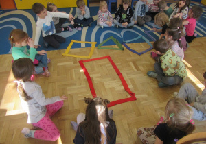 Dzieci siedzą w kole, przed nimi figury geometryczne ułożone z szarf.