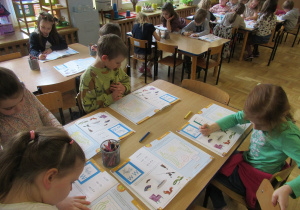 Dzieci siedzą przy stolikach i pracują w książkach.