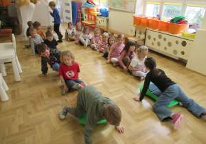 Dzieci siedzą w dwóch rzędach, dwoje dzieci kładzie się na jeździkach gimnastycznych.