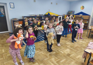 Dzieci stoją w sali i trzymają w rękach przytulanki.