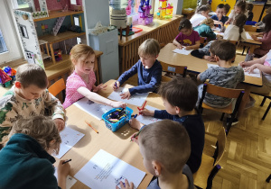 Dzieci siedzą przy stoliku i wykonują karty pracy.