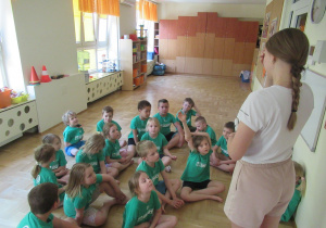 Dzieci w strojach gimnastycznych siedzą na podłodze podczas zajęć z elementami taekwondo.