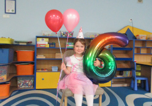 Dziewczynka obchodzi urodziny, siedzi na krześle i trzyma w rękach balony.