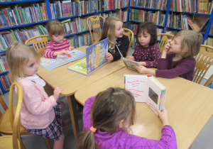 Dziewczynki siedzą przy stoliku w bibliotece i oglądają książki.