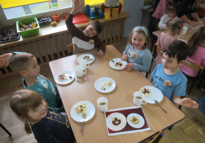 Dzieci podczas misiowego podwieczorku - dzieci mają na talerzach ułożone z wafla ryżowego, marmolady, kawałków banana i rodzynek buzie misia.