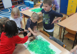 Dzieci stoją przy stolikach i malują choinki na dużych kartkach.