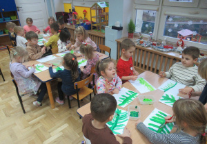 Dzieci siedzą przy stolikach i malują farbami gałązki choinkowe.