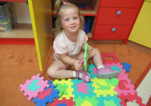Dziewczynka siedzi na dywanie i układa piankowe puzzle.