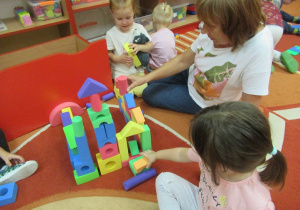 Dzieci i Pani siedzą na dywanie i budują zamek z klocków.