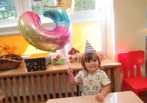 Dziewczynka siedzi przy stoliku i trzyma balon z cyfrą 3.