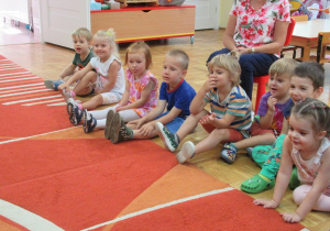 Przedszkolaki siedzą na dywanie i biorą udział w zajęciach.