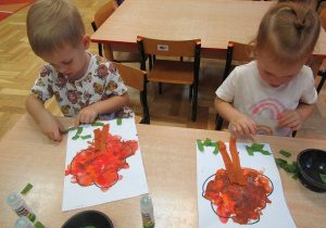 Dzieci siedzą przy stolikach i wyklejają drzewo kolorowym papierem.