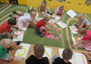 Dzieci siedzą na dywanie i układają kształty z kolorowych nakrętek.