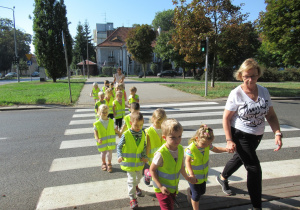 Dzieci w odblaskowych kamizelkach przechodzą przez przejście dla pieszych.