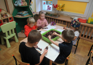 Dzieci siedzą przy stoliku i układają kształty z kasztanów.