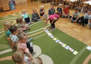Dzieci siedzą w kole na dywanie, dziewczynka układa obrazki.
