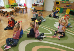 Dzieci siedzą w parach plecami do siebie i podają sobie nad głowami woreczki gimnastyczne.