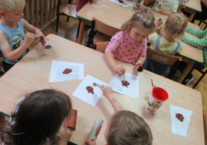 Dzieci przy stolikach malują rolki po papierze brązową farbą.