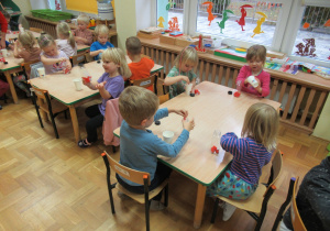Dzieci siedzą przy stolikach i wrzucają pomponiki do butelek.