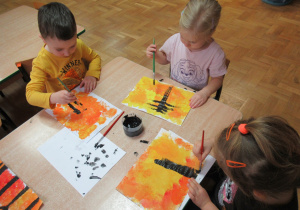 Dzieci siedzą przy stole i malują drzewa farbami.