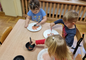 Dzieci siedzą przy stolikach i malują papierowe talerzyki na kolor brązowy.