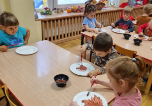 Przedszkolaki siedzą przy stolikach i malują papierowe talerzyki brązową farbą.