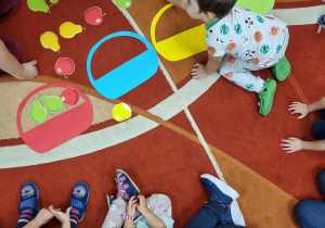 Dzieci siedzą na dywanie i układają obrazki z koszyczkami i owocami.