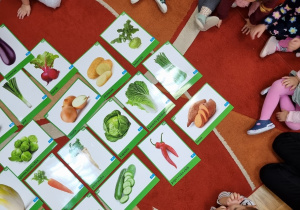 Dzieci siedzą na dywanie i oglądają ilustracje z warzywami.