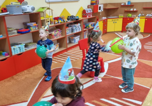 Dzieci biegają po czerwonym dywanie z balonikami i czapeczkami na głowie.