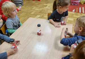 Dzieci siedzą przy stolikach i trzymają butelki z biało-czerwonymi pomponami.