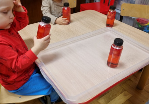 Dzieci siedzą przy stoliku i trzymają butelki z czerwonym płynem.