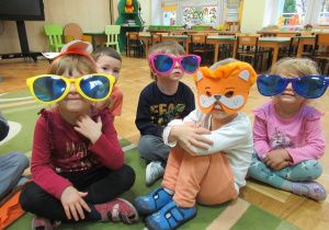 Dzieci siedzą na dywanie, chłopiec w masce lwa i dzieci w dużych okularach.