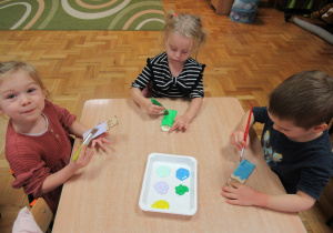 Dzieci przy stoliku malują farbami zakładki do książki.
