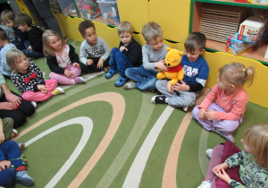 Dzieci siedzą w kole i podaja sobie misia.