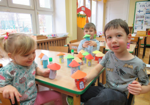 Dzieci siedzą przy stoliku i prezentują prace plastyczne - domki Muminków.