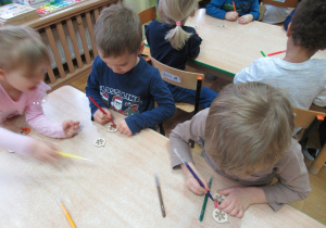 Dzieci siedzą przy stolikach i kolorują flamastrami drewniane bombki.