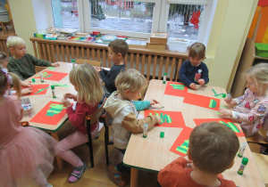 Dzieci siedzą przy stolikach i przyklejają zielone paski na kartki.
