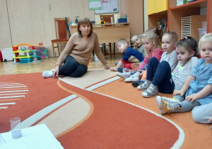Dzieci siedzą na dywanie i biorą udział w zajęciach.