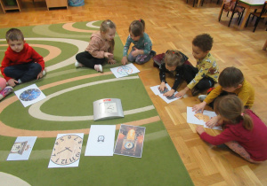 Ddzieci siedzą na dywanie, przed nimi obrazki przedstawiające zegary.
