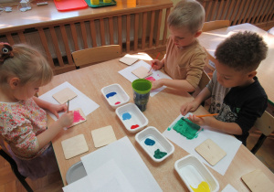 Dzieci przy stoliku malują farbami drewniane podkładki.