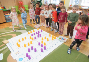 Dzieci podczas zajęć z kodowania - dzieci stoją wokół maty, na której ułożone są kolorowe kubeczki.