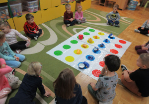 Dzieci siedzą w kole na dywanie, przed nimi mata z kolorowymi figurami.