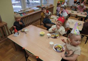 Dzieci siedzą przy stolikach, zjadają urodzinowy poczęstunek.