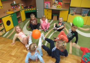 Dzieci siedzą w kole i podają sobie balon stopami.