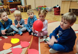 Dzieci siedzą na dywanie, na którym leżą kolorowe, papierowe baloniki.i