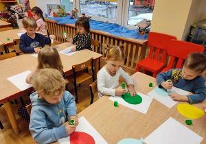 Dzieci siedzą przy stolikach i naklejają kolorowe, papierowe baloniki na białą kartkę.