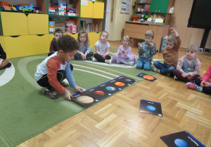 Dzieci siedzą na podłodze podzas zajęć, chłopiec układa obrazki przedstawiające planety.