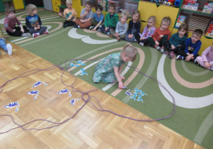 Dzieci siedzą na dywanie, dziewczynka układa obrazki dinozaurów.